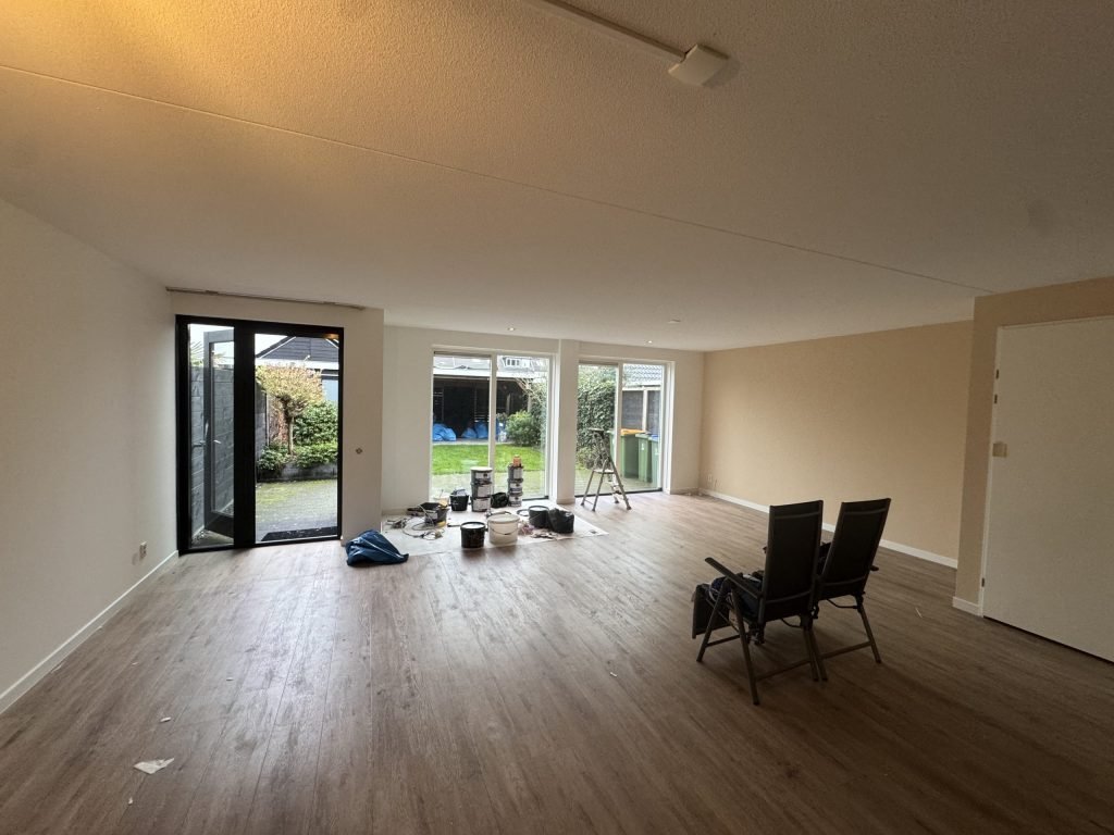 Woonkamer van een bestaande woning met tuindeuren, een houten vloer en verfspullen op een afdekkleed