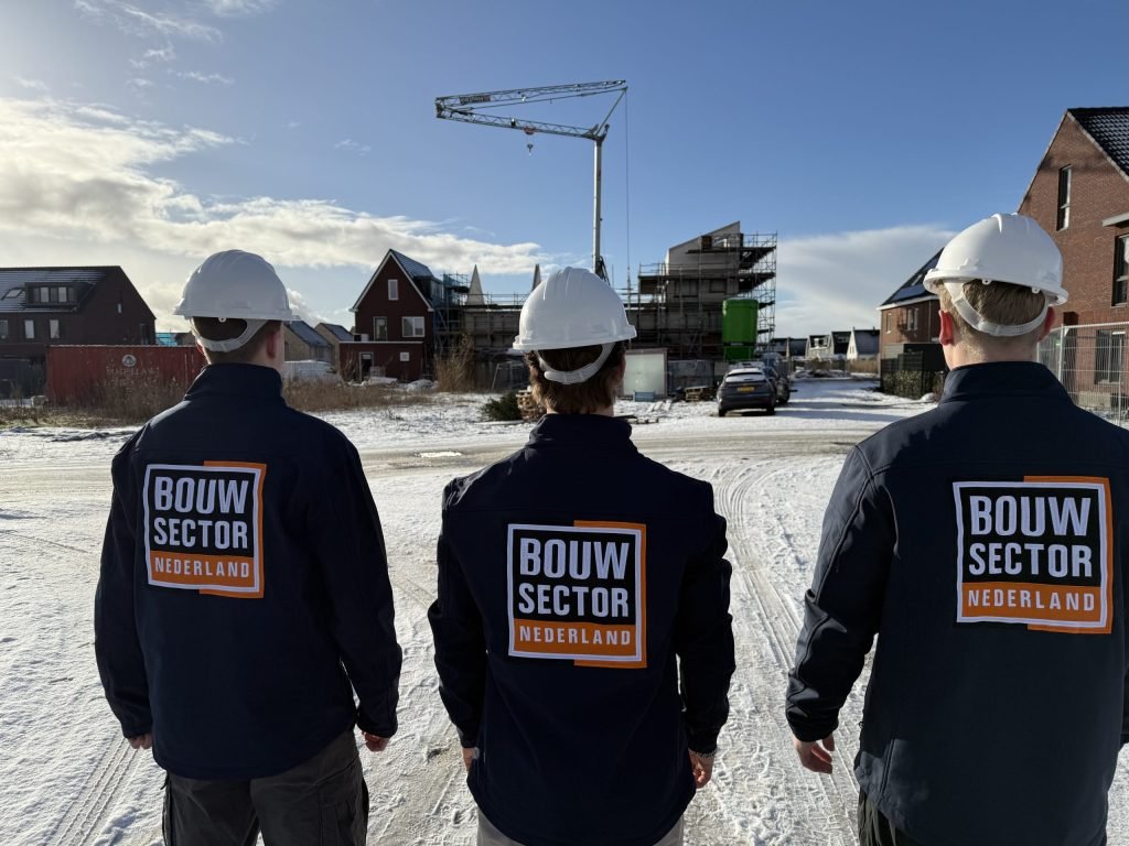 Drie vakmensen van Bouwsector Nederland in bedrijfsjas en witte helm kijken naar een nieuwbouwwijk in de sneeuw
